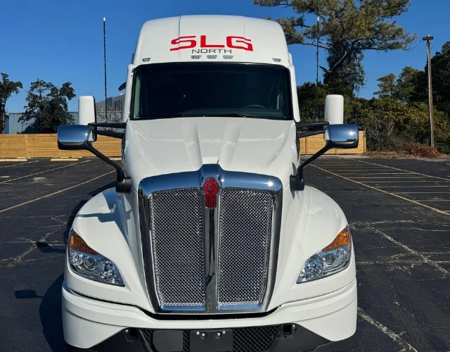 White semi-truck with "SLG North" on cab, parked on a sunny day in a lot with trees and wooden fence in the background.